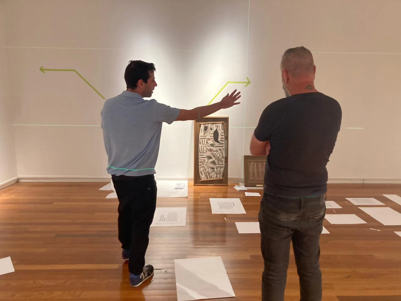 Two people stand in the gallery during installation, one gesturing at arrows taped to the wall, with label texts laid out on the floor