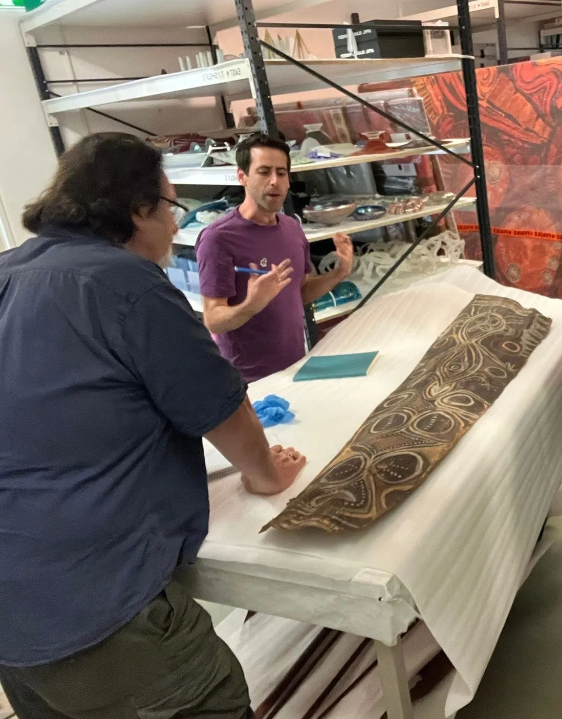 A person in a purple shirt studies the painted panel laid on a table beneath storage shelves
