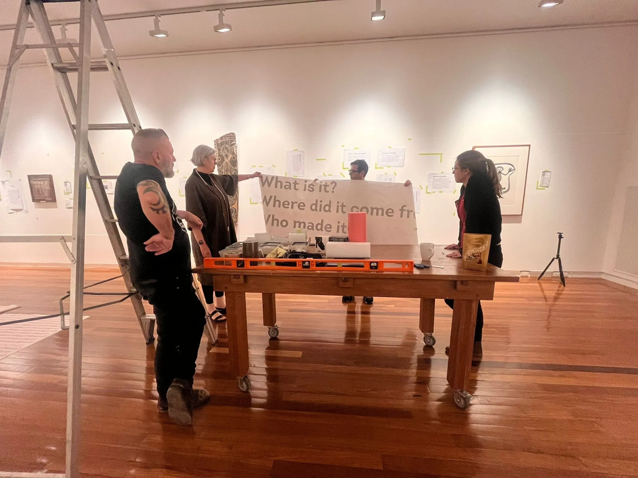 Four people around a table in the gallery discussing the hang, with a ladder and a pin board of the four questions behind them