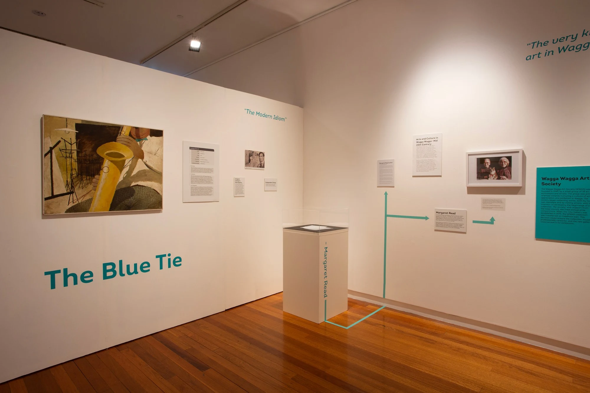 The Blue Tie on the first wall, its title painted beneath it in teal, with a plinth and teal lines leading to Margaret Read